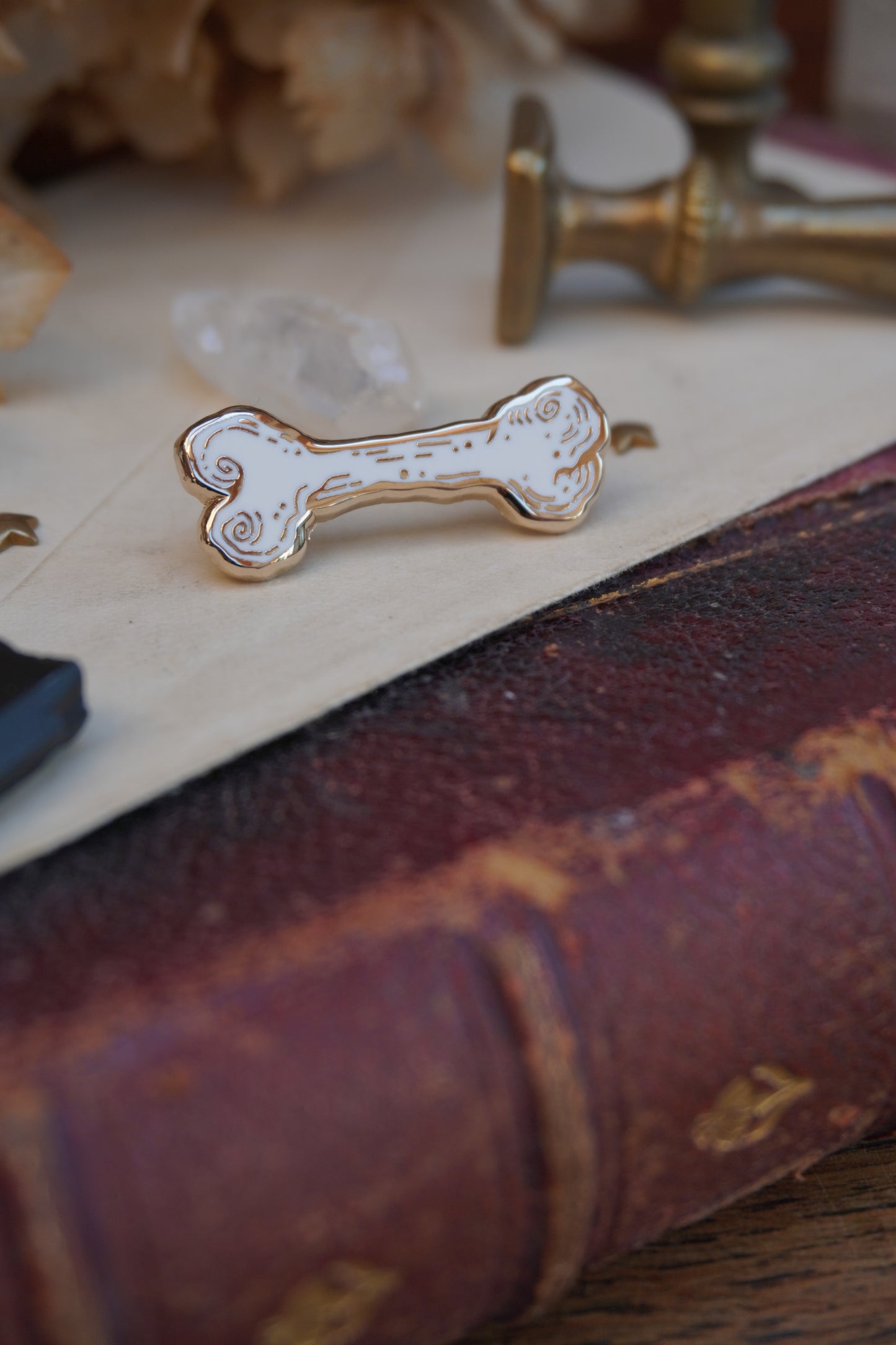 A close up of a bone shaped enamel pin in white and gold, resting on a small white crystal displayed on vintage paper on an old book with dried flowers and wax seals in the background