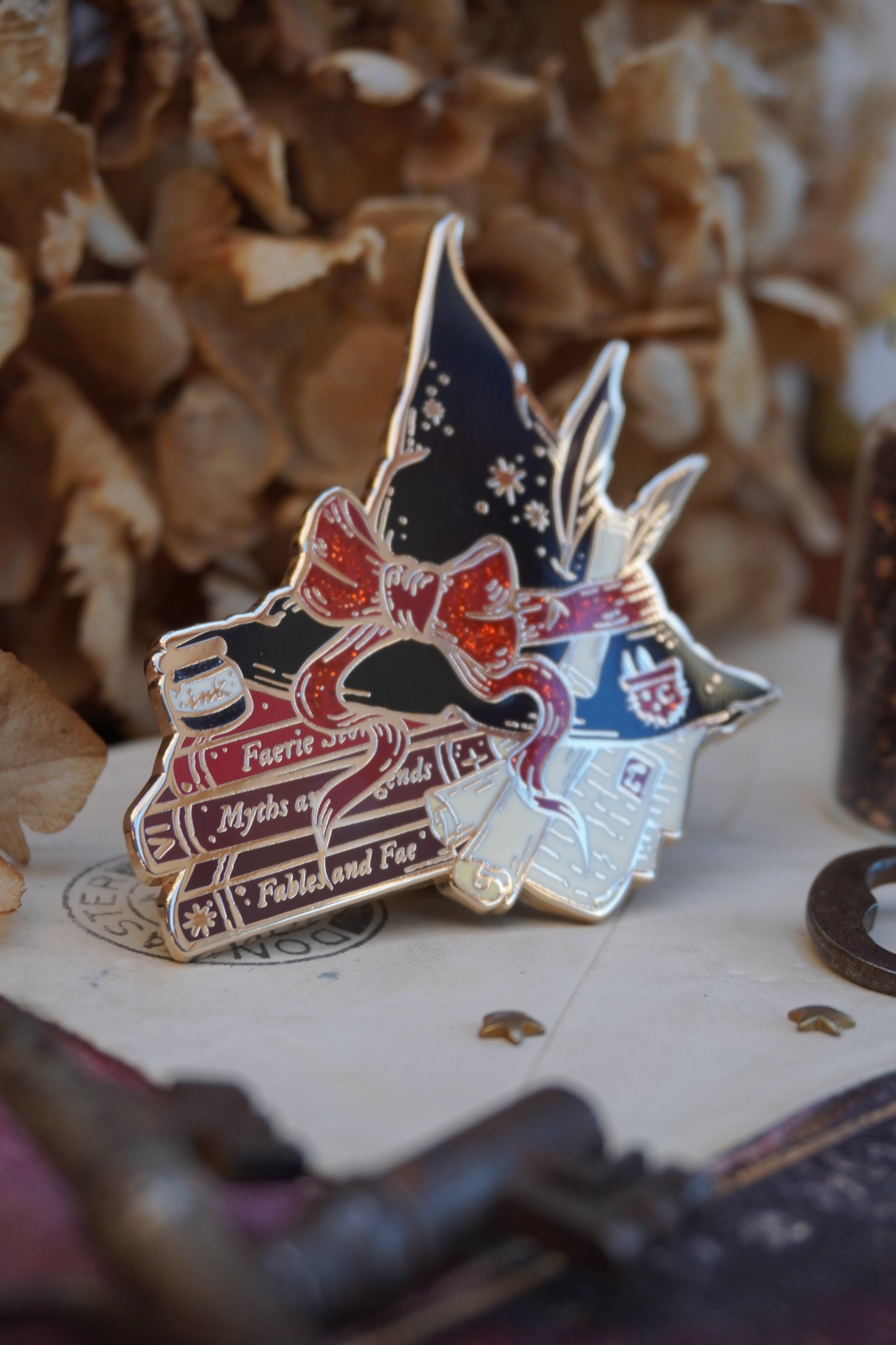 An enamel pin shaped into a witch hat on top of books and scrolls, resting on a vintage style surface with dried flowers in the background.