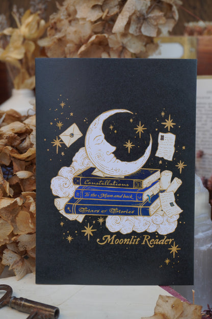 A black postcard with celestial design with the text  'Moonlit Reader' stood on top of vintage paper with a decorative background with dried flowers