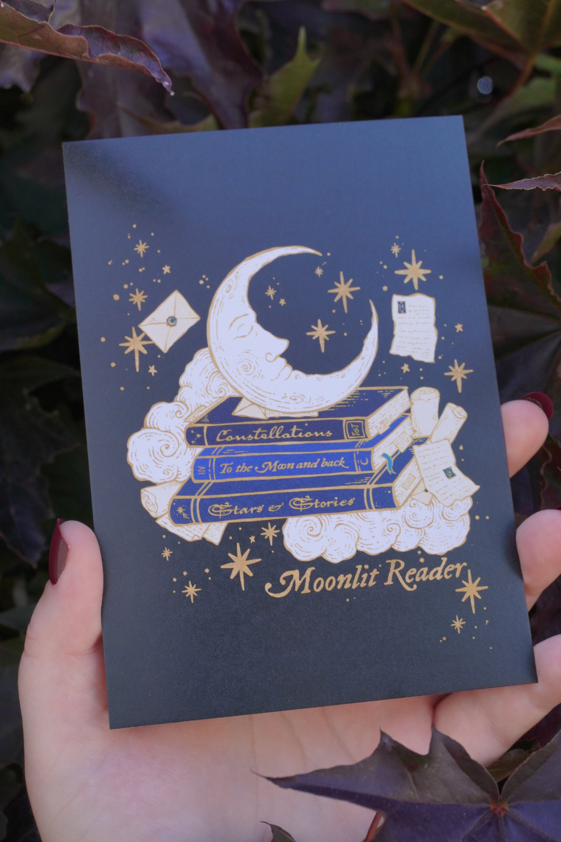 Hand holding a black postcard with a gold white and blue. crescent moon, stars, clouds, books, and scrolls decoration with the text  'Moonlit Reader' aginst dark purple leaves