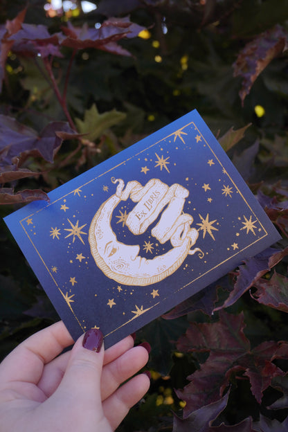 A hand holding a blue toned postcard featuring gold-toned celestial illustration of a crescent moon, stars, and a central banner reading 'Ex Libris'. Set against a background of leaves.