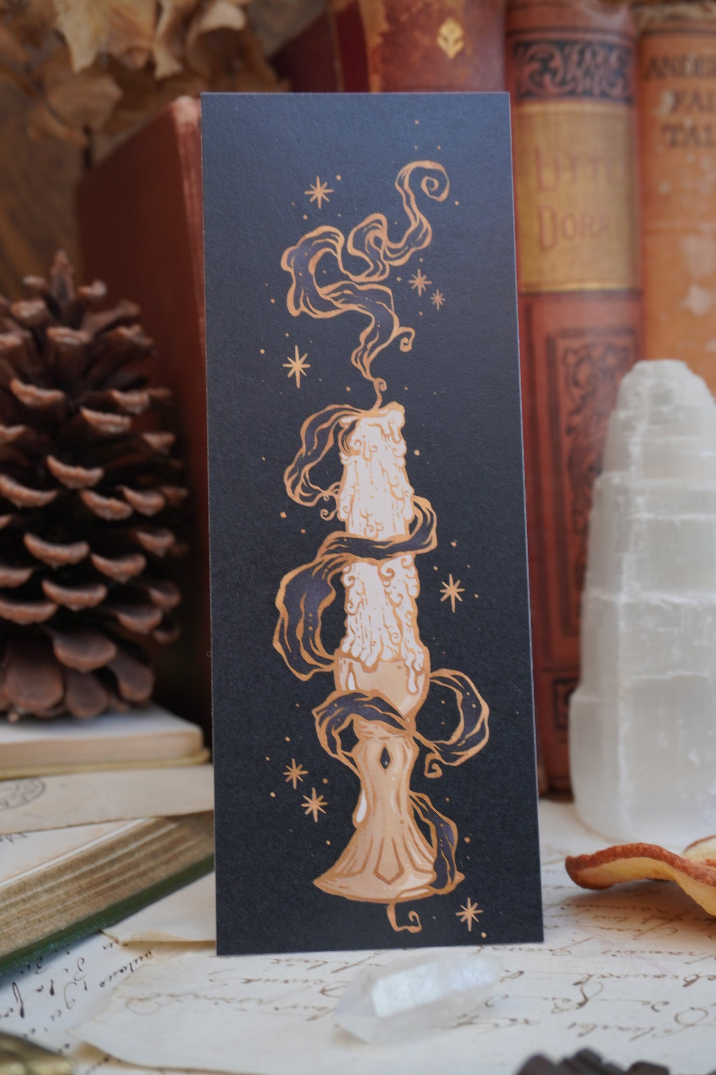 A black bookmark featuring a detailed illustration of a melting candle with gold stars and swirls and grey smoke, displayed on top of vintage books and a pinecone