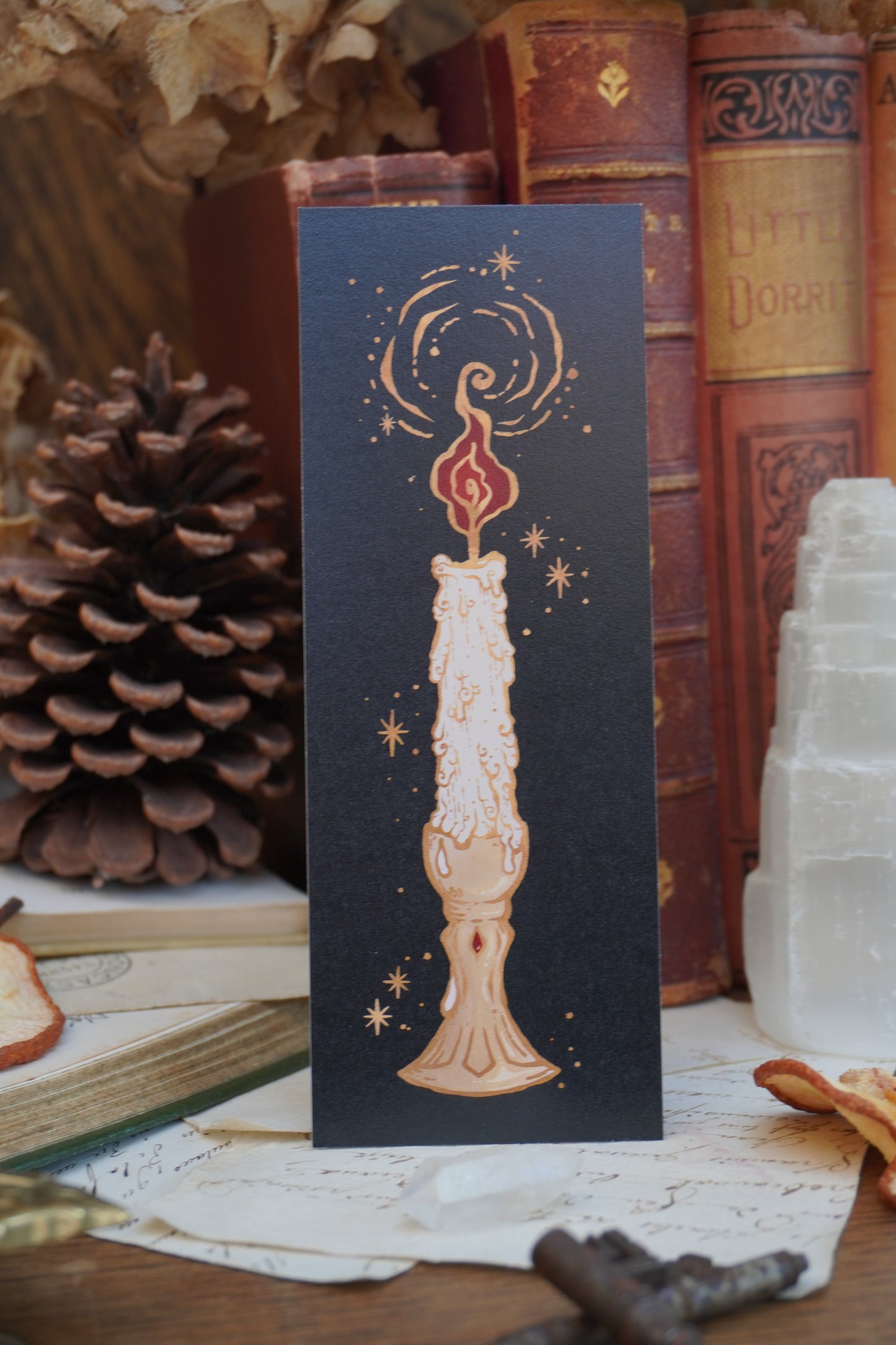 A black bookmark featuring a detailed illustration of a melting candle with gold stars and swirls, displayed on top of vintage books and a pinecone