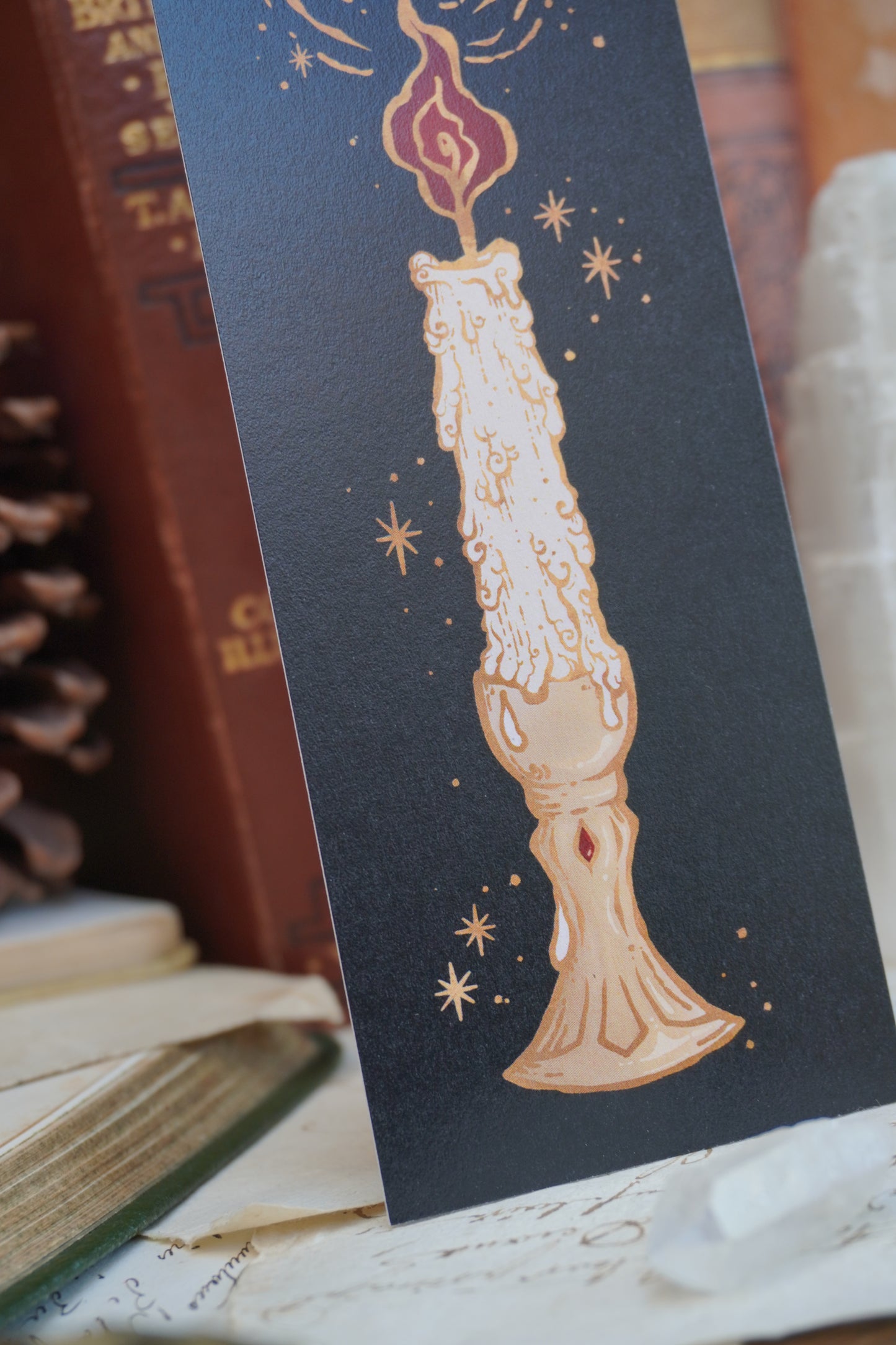 a close up of a  black bookmark featuring a detailed illustration of a melting candle with gold stars and swirls, displayed on top of vintage books and a pinecone