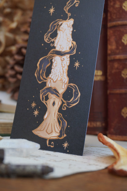 a close up of a black bookmark featuring a detailed illustration of a melting candle with gold stars and swirls and grey smoke, displayed on top of vintage books and a pinecone