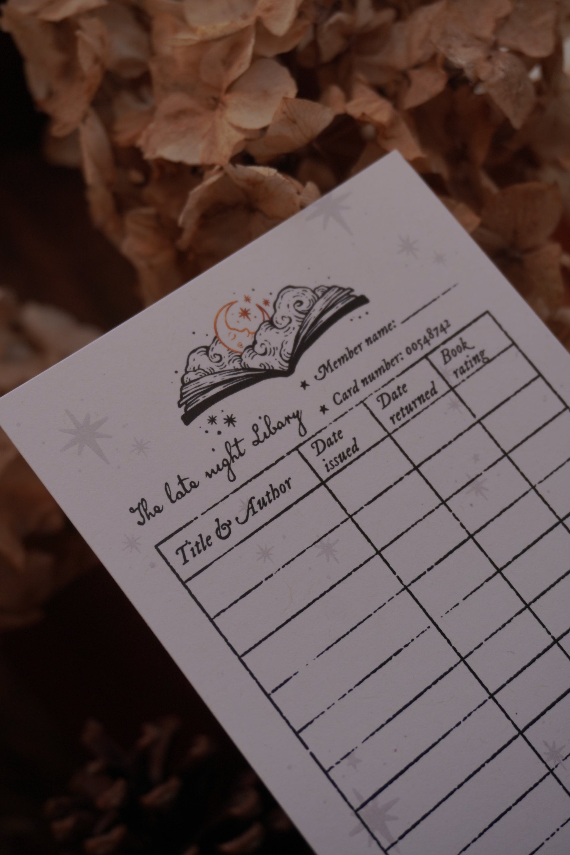 A close-up of a printed library card bookmark 'The late night libary' with fields for member name, card number, title and author, date issued, date returned, and book rating. Photographed on a dark background with dried beige flowers.