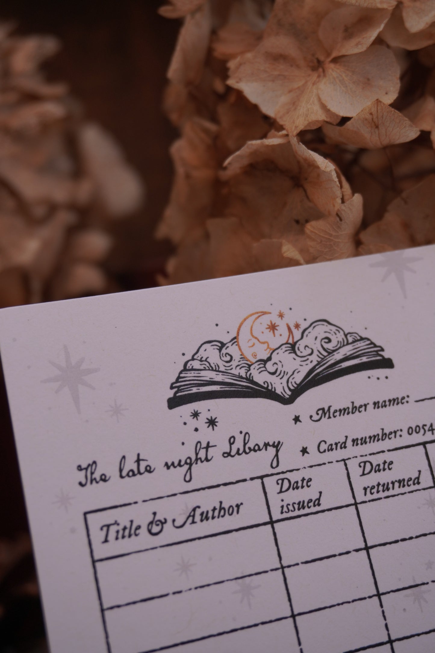 A close-up of a white paper library card motif bookmark with a decorative open-book illustration with text underneath placed against dried beige flowers.