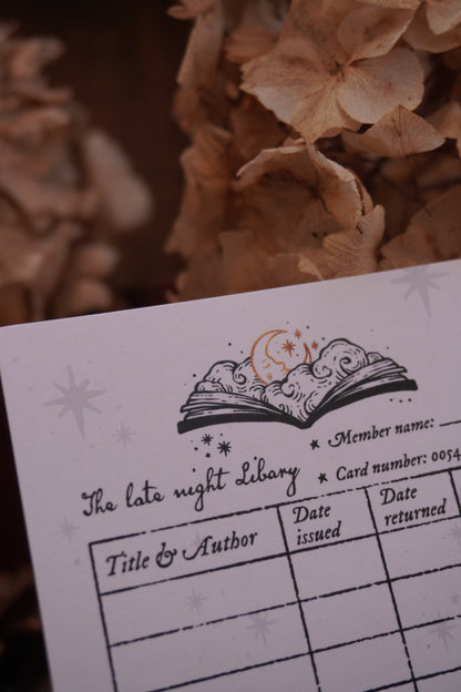 A close-up of a white paper library card motif bookmark with a decorative open-book illustration with text underneath placed against dried beige flowers.