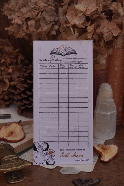 A white paper library card motif bookmark with celestial illustrations, styled on a wooden surface surrounded by dried flowers, crystals, and pinecones.
