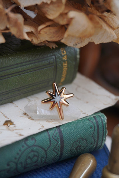 A gold star enamel pin with a pearl swirl star shaped centre, resting on a crystal on a stack of vintage green books with vintage paper and dried flowers in the background.