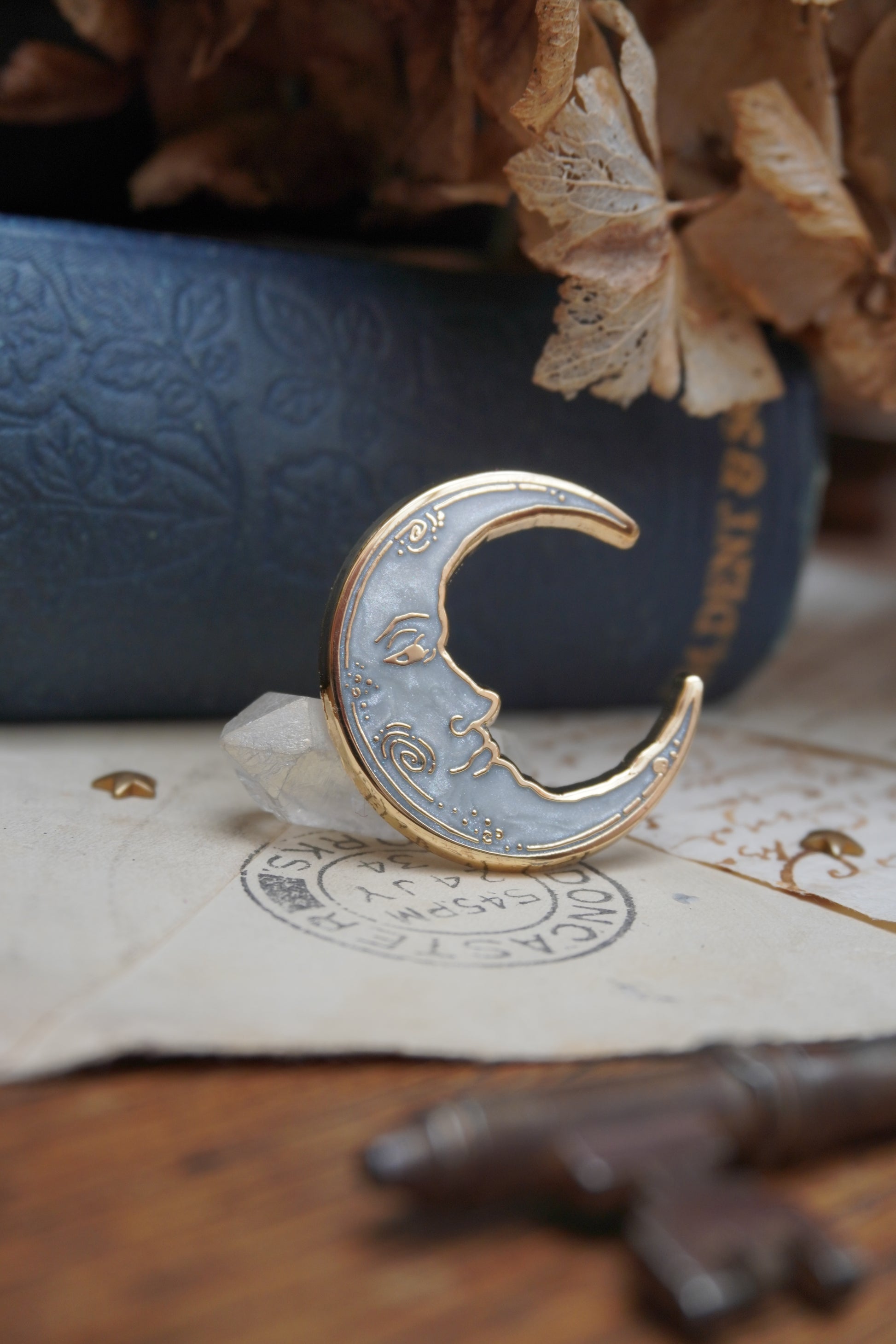 A vintage man in the moon, crescent shaped enamel pin with white pearl swirl enamel and gold details rests on vintage paper with a decorative background of a blue vintage book and dried flowers