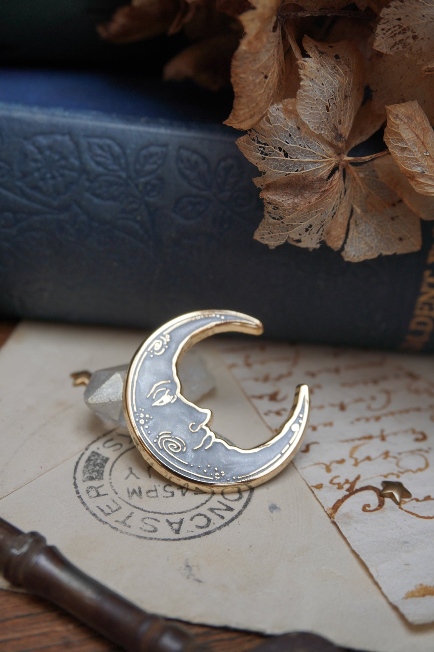 A vintage man in the moon, crescent shaped enamel pin with white pearl swirl enamel and gold details rests on a small crystal and vintage paper with a decorative background of a blue vintage book and dried flowers