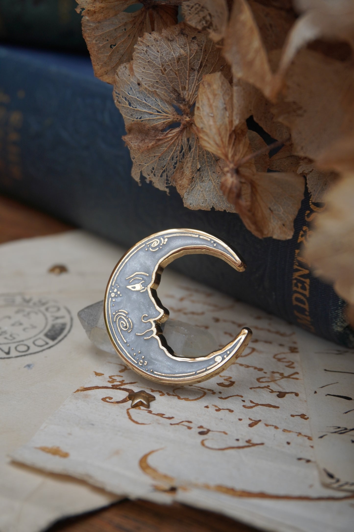 A vintage man in the moon, crescent shaped enamel pin with white pearl swirl enamel and gold details rests on vintage paper with a decorative background of a blue vintage book and dried flowers
