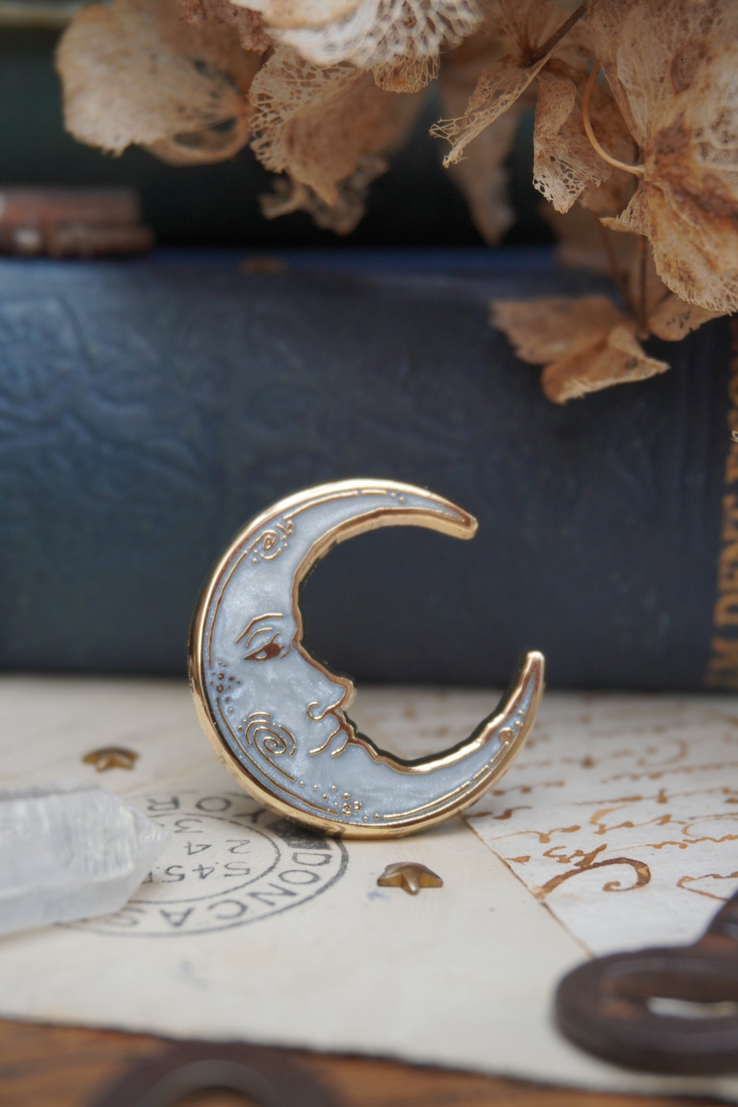 A vintage man in the moon, crescent shaped enamel pin with white pearl swirl enamel and gold details rests on vintage paper, shown close up against vintage-style books and dried flowers.