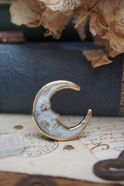 A vintage man in the moon, crescent shaped enamel pin with white pearl swirl enamel and gold details rests on vintage paper, shown close up against vintage-style books and dried flowers.