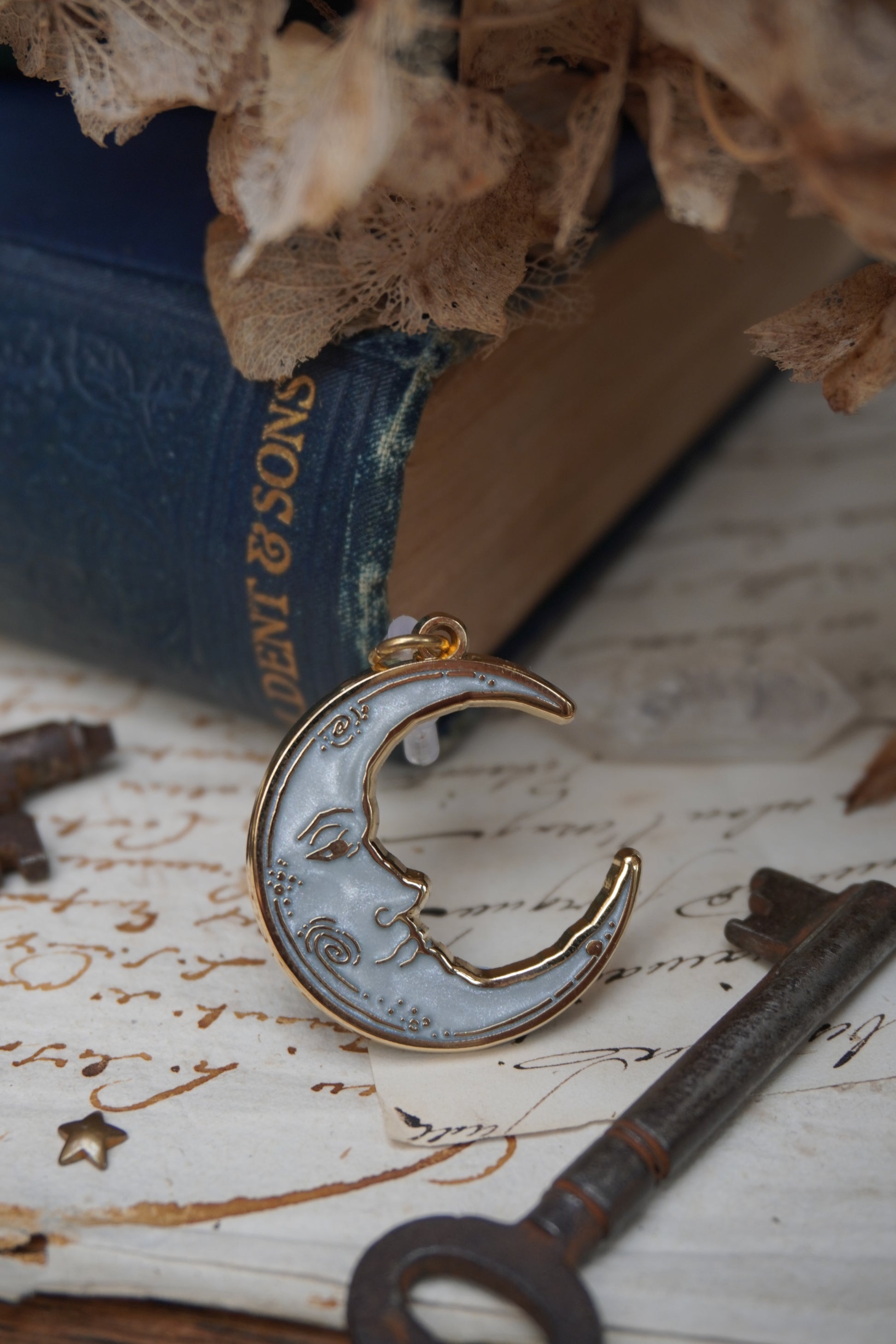 A vintage man in the moon, crescent shaped dust plug with white pearl swirl enamel and gold details rests on vintage paper with a decorative background of vintage books, dried flowers and keys.