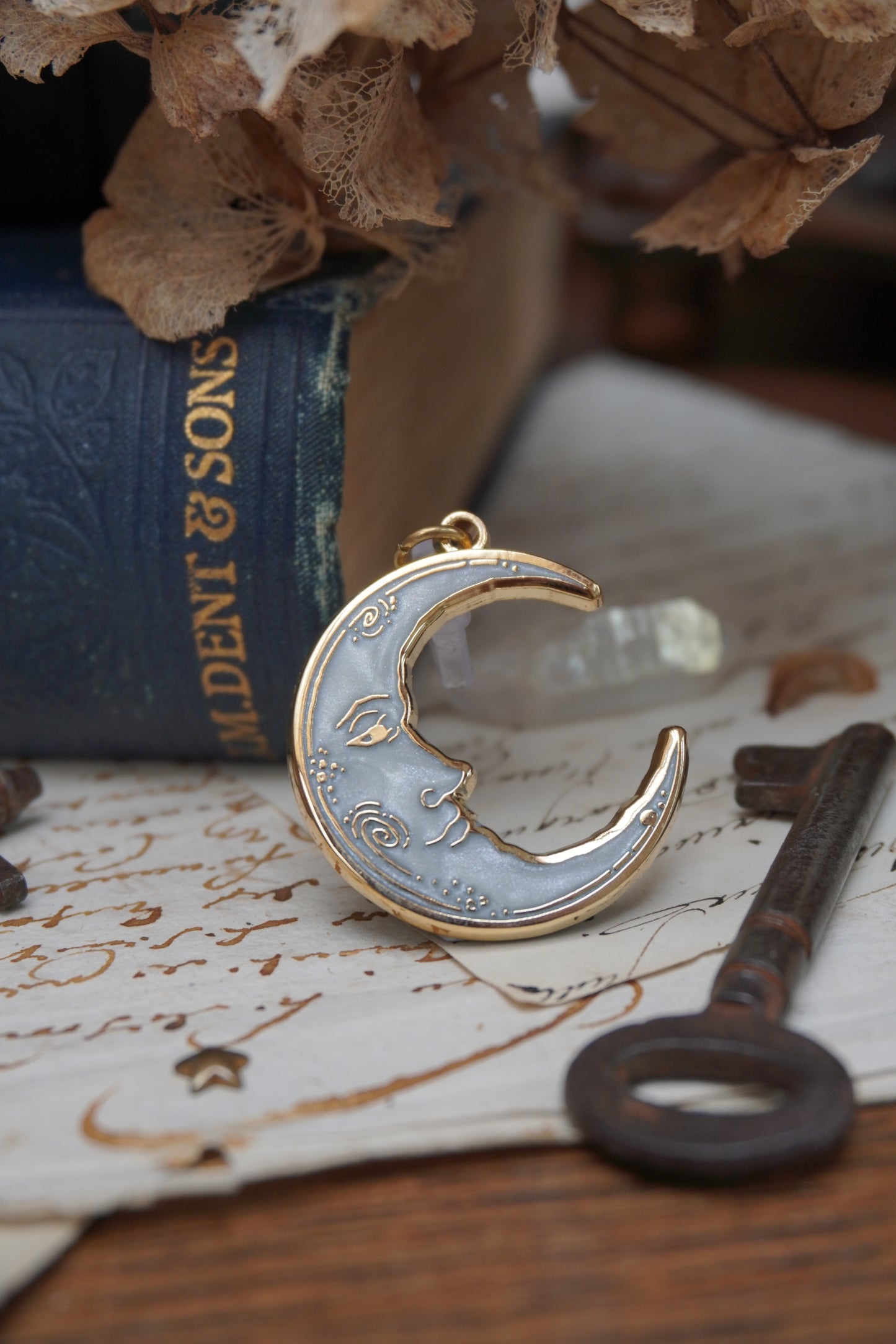 A vintage man in the moon, crescent shaped dust plug with white pearl swirl enamel and gold details rests on vintage paper with a decorative background of vintage books, dried flowers and keys with a small crystal 