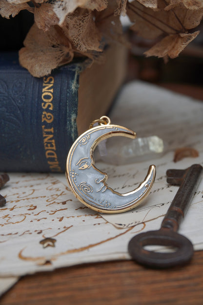 A vintage man in the moon, crescent shaped dust plug with white pearl swirl enamel and gold details rests on vintage paper with a decorative background of vintage books, dried flowers and keys with a small crystal 