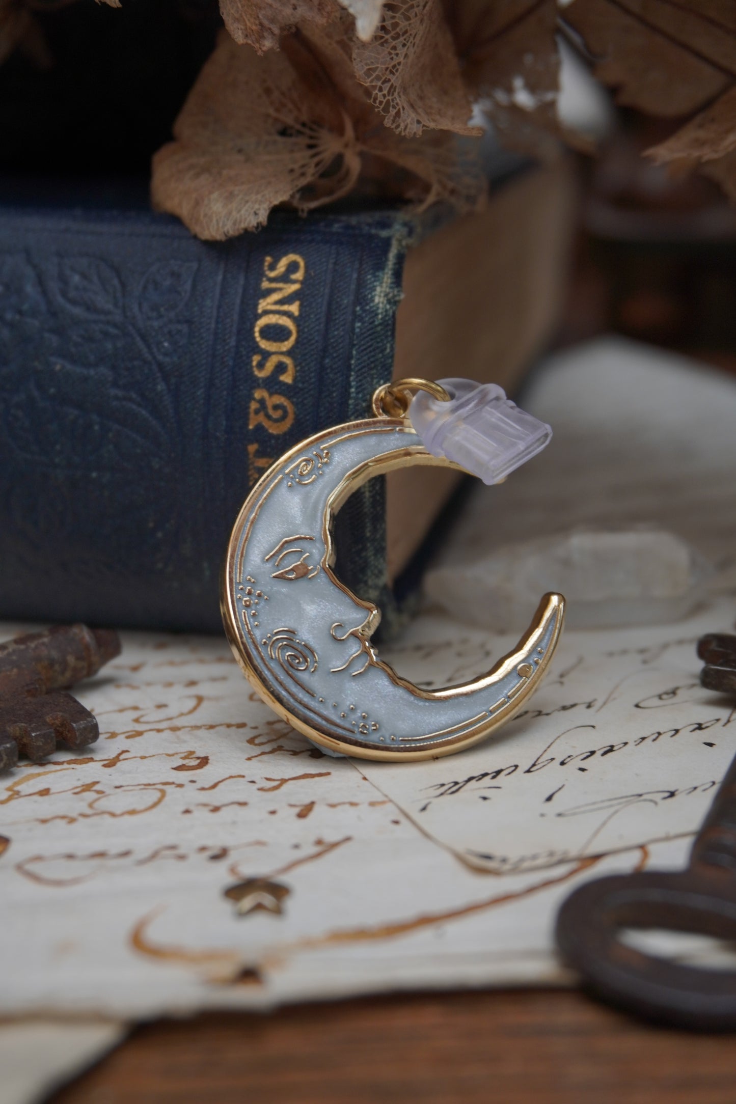 A vintage man in the moon, crescent shaped dust plug with clear plastic USB C attachment; with white pearl swirl enamel and gold details rests on vintage paper with a decorative background of vintage books, dried flowers and keys.