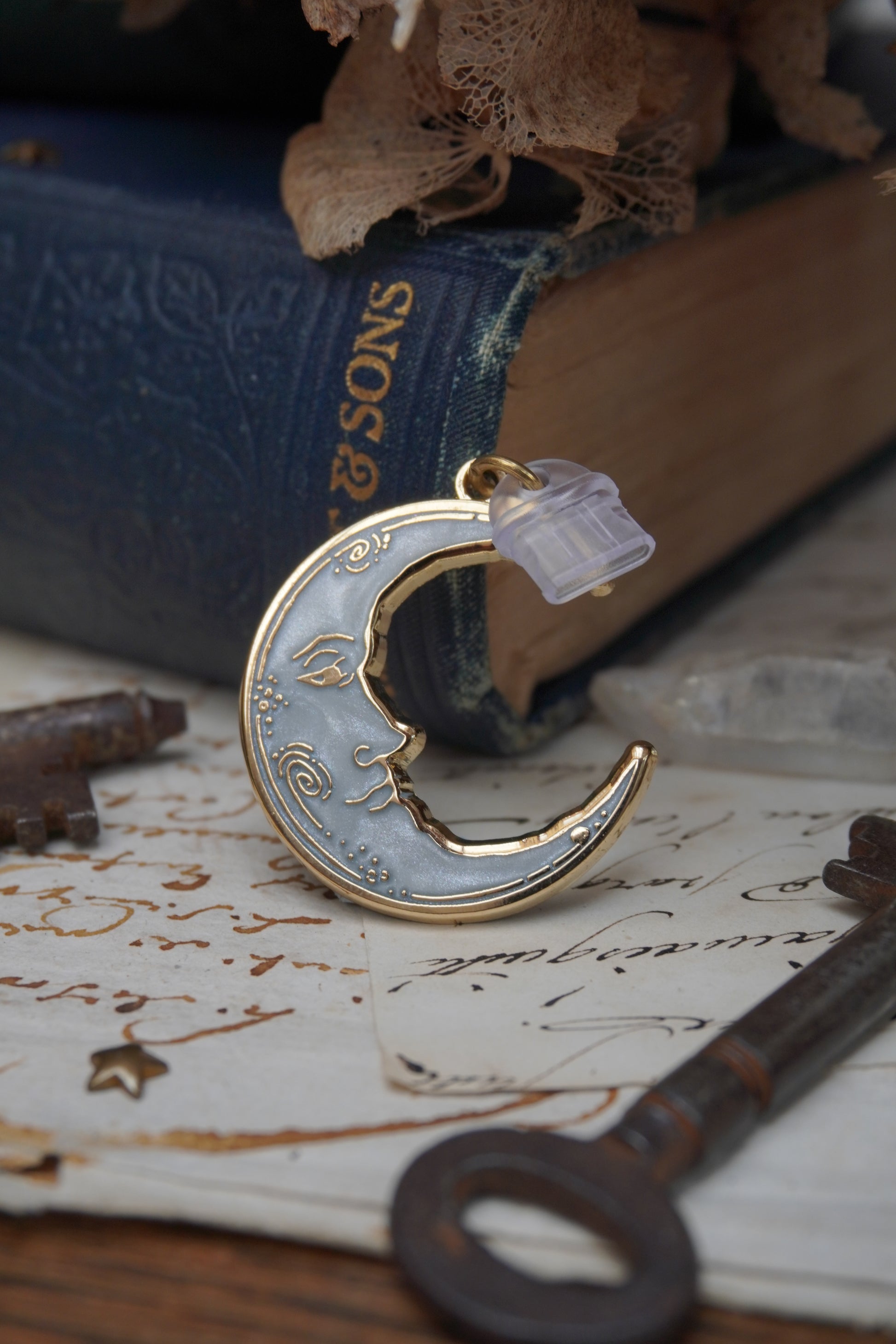 A vintage man in the moon, crescent shaped dust plug with white pearl swirl enamel and gold details rests on vintage paper with a decorative background of vintage books, dried flowers and keys.