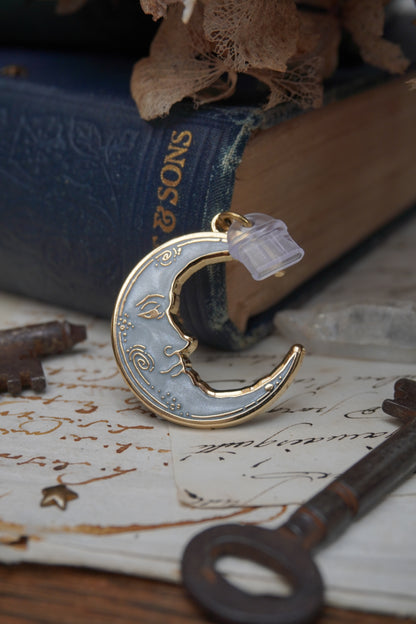 A vintage man in the moon, crescent shaped dust plug with white pearl swirl enamel and gold details rests on vintage paper with a decorative background of vintage books, dried flowers and keys.