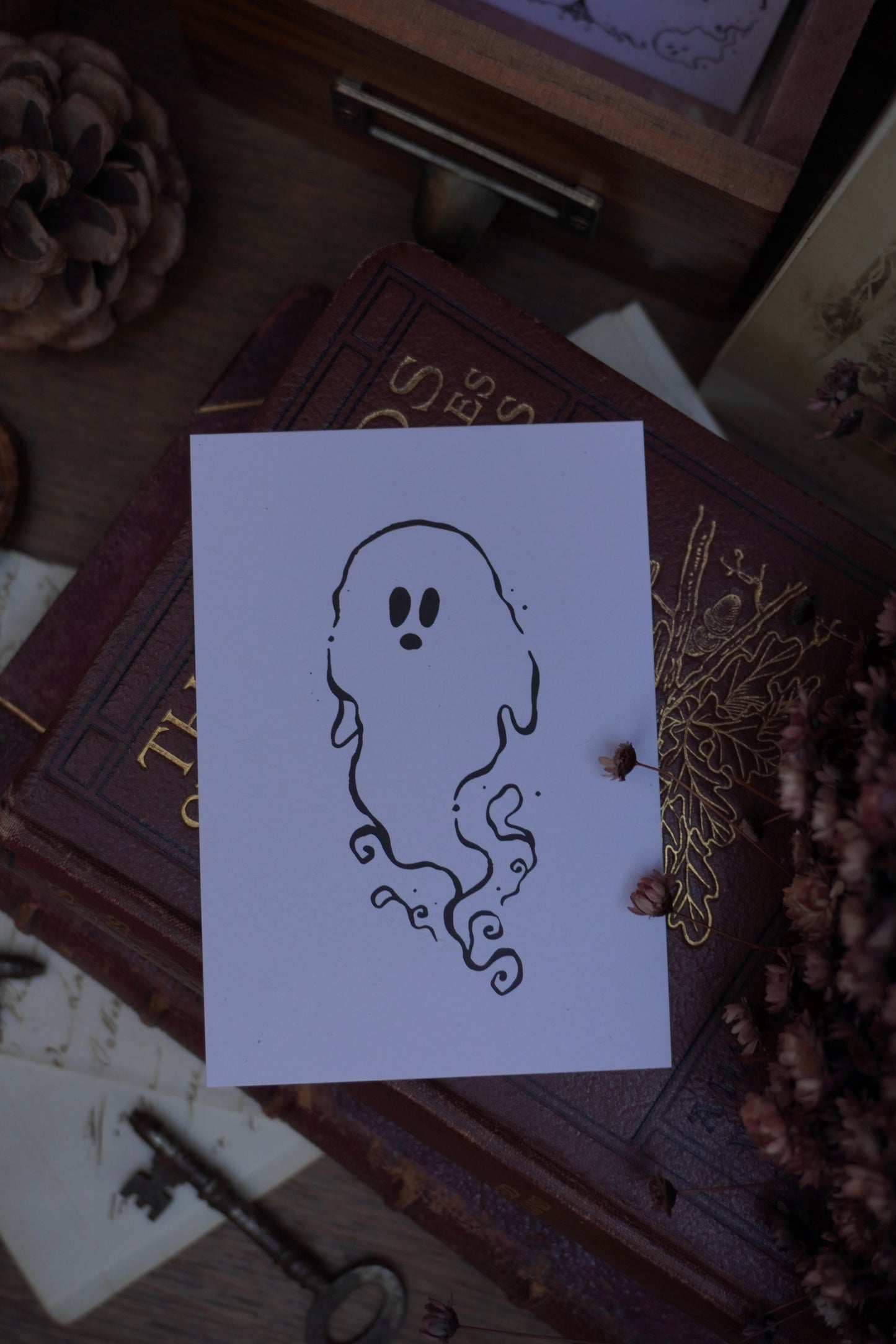 Swirly black outline ghost drawing on white card, resting on a vintage book surrounded by decorative items like dried flowers, vintage paper and a key in the background