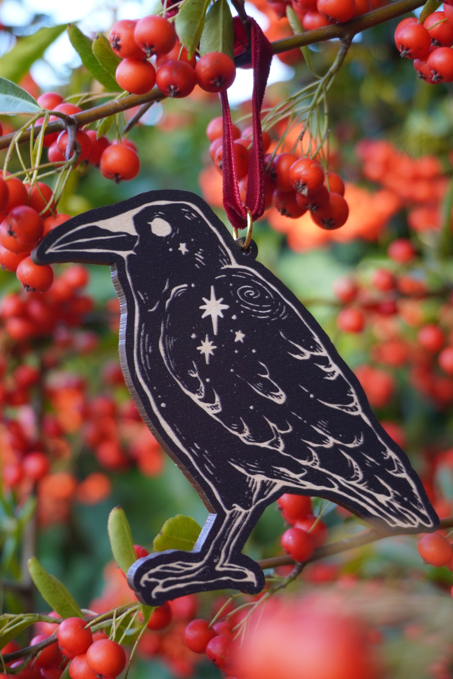 Wooden hanging ornament in the shape of a crow with a crow and stars design, resting on a red berry bush