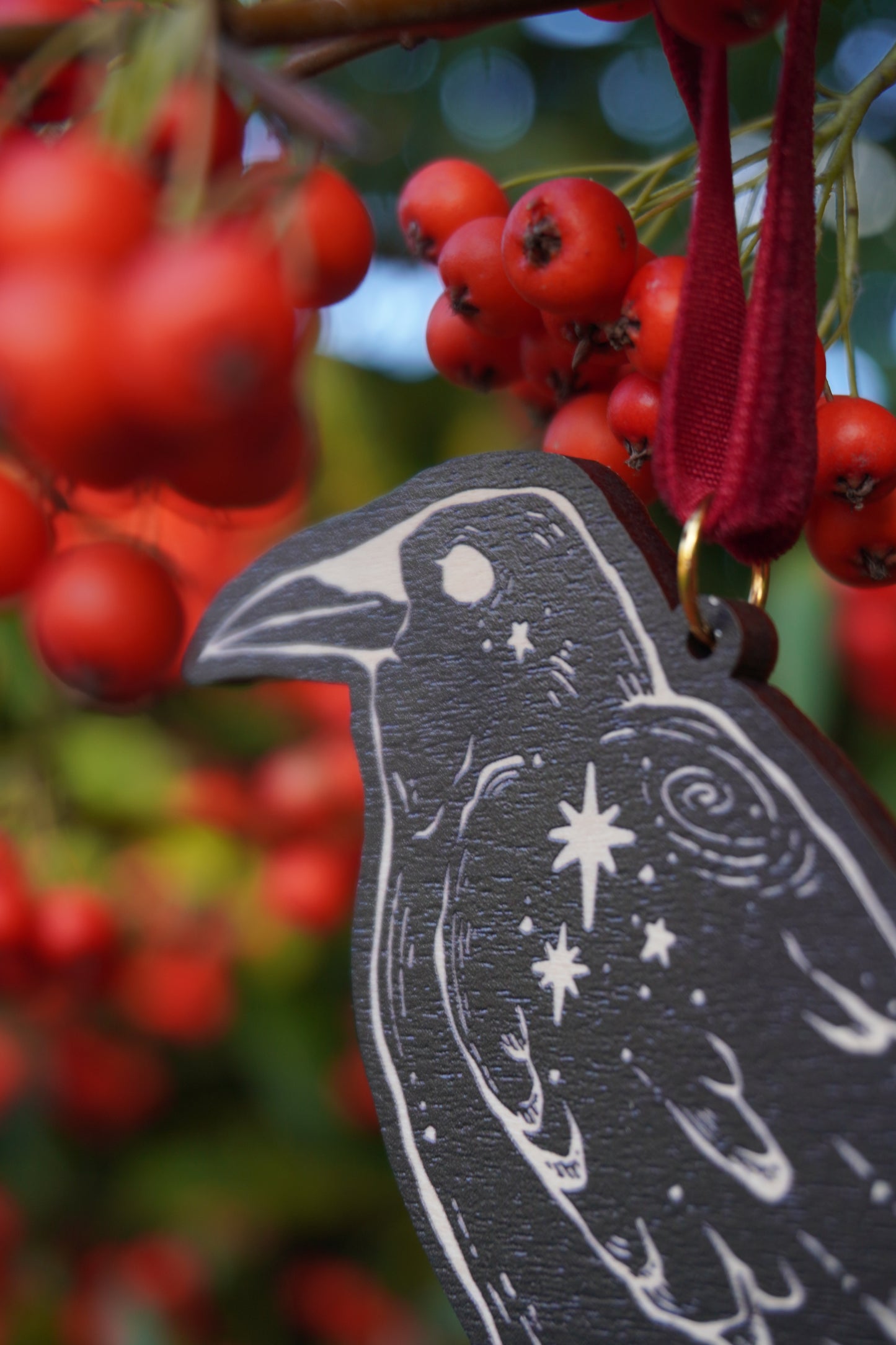 Wooden hanging ornament in the shape of a crow with a crow and stars design, resting on a red berry bush
