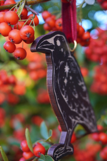 Wooden hanging ornament in the shape of a crow with a crow and stars design hanging on a red berry bush 