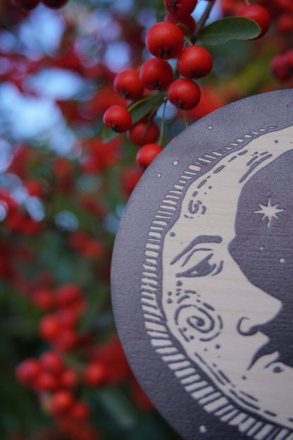Circular wooden hanging ornament with a celestial moon and stars design with a red berry bush background