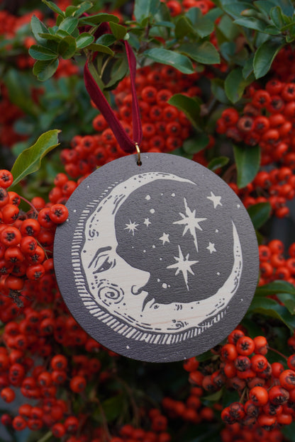 Circular wooden hanging ornament with a celestial moon and stars design, resting on a red berry bush