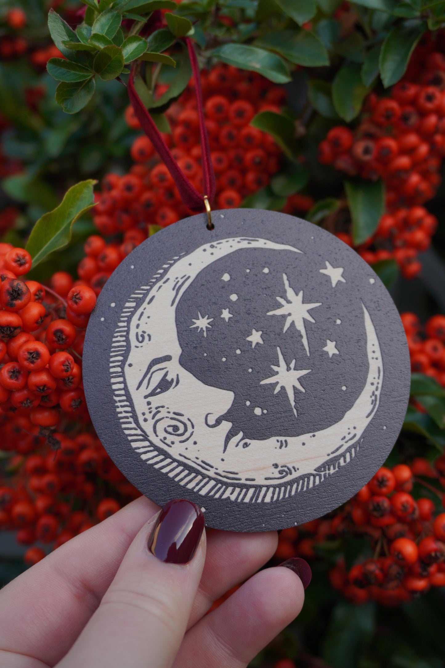 Hand holding a circular wooden hanging ornament with a celestial moon and stars design, resting on a red berry bush