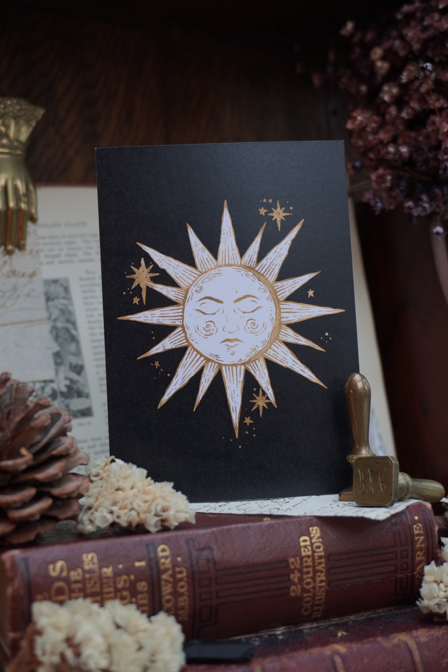 Postcard with a sun design in white and gold on a black background, resting on a wooden table with books and dried flowers