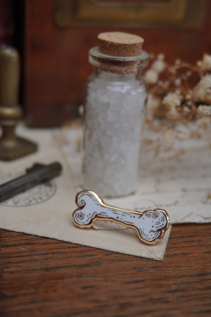 A bone shaped enamel pin in white and gold, resting on a small white crystal displayed on vintage paper on a wooden surface, next to a glass bottle with a cork stopper, an old key and dried flowers