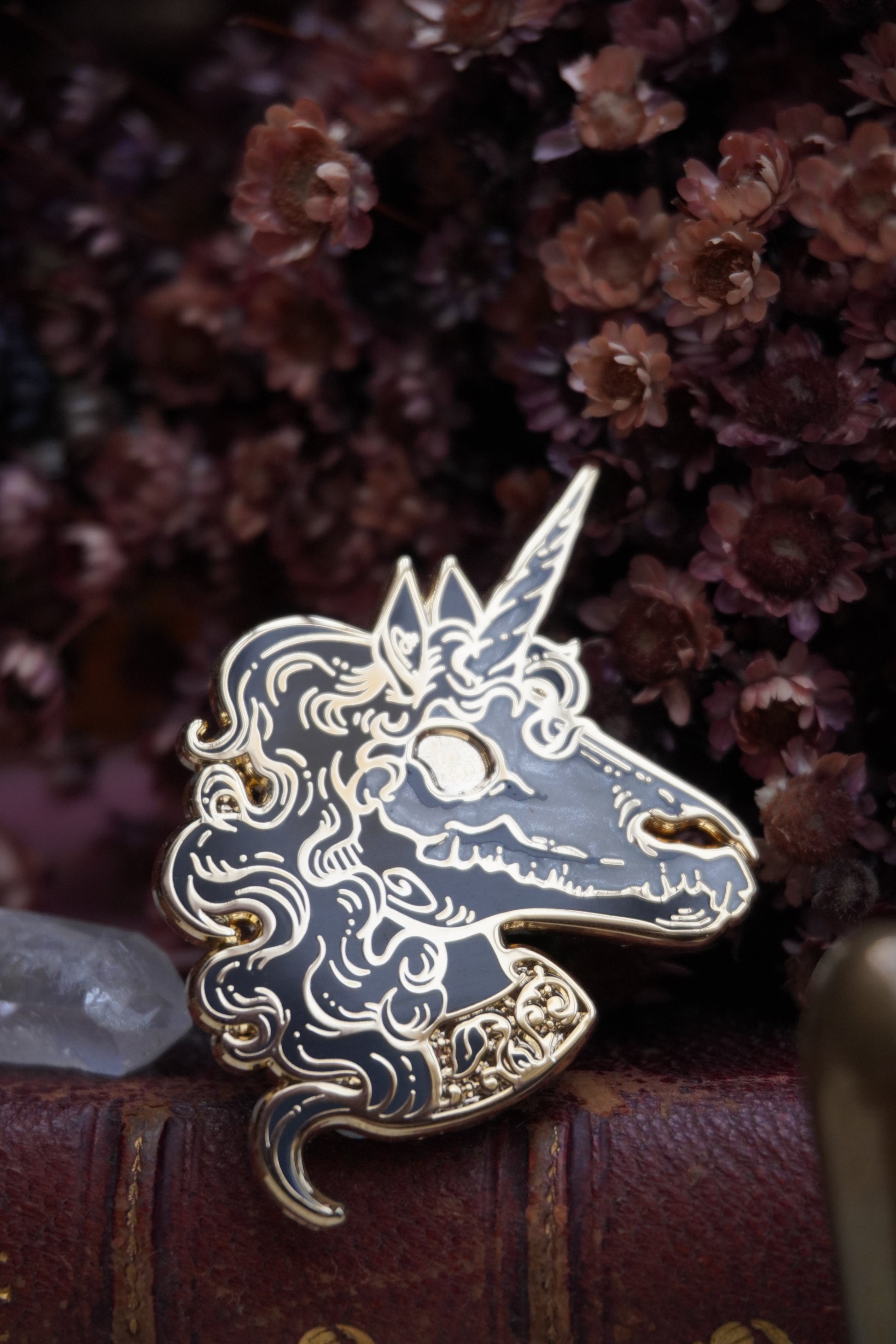 A close up of an enamel pin shaped like a unicorn head. The pin features a black face and neck with white pearl effect skull on top of the face, grey curly mane, lighter grey screen print details and gold plated metal outlines resting on vintage paper with dried dark red flowers and a crystal in the background 