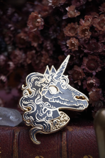 A close up of an enamel pin shaped like a unicorn head. The pin features a black face and neck with white pearl effect skull on top of the face, grey curly mane, lighter grey screen print details and gold plated metal outlines resting on vintage paper with dried dark red flowers and a crystal in the background 