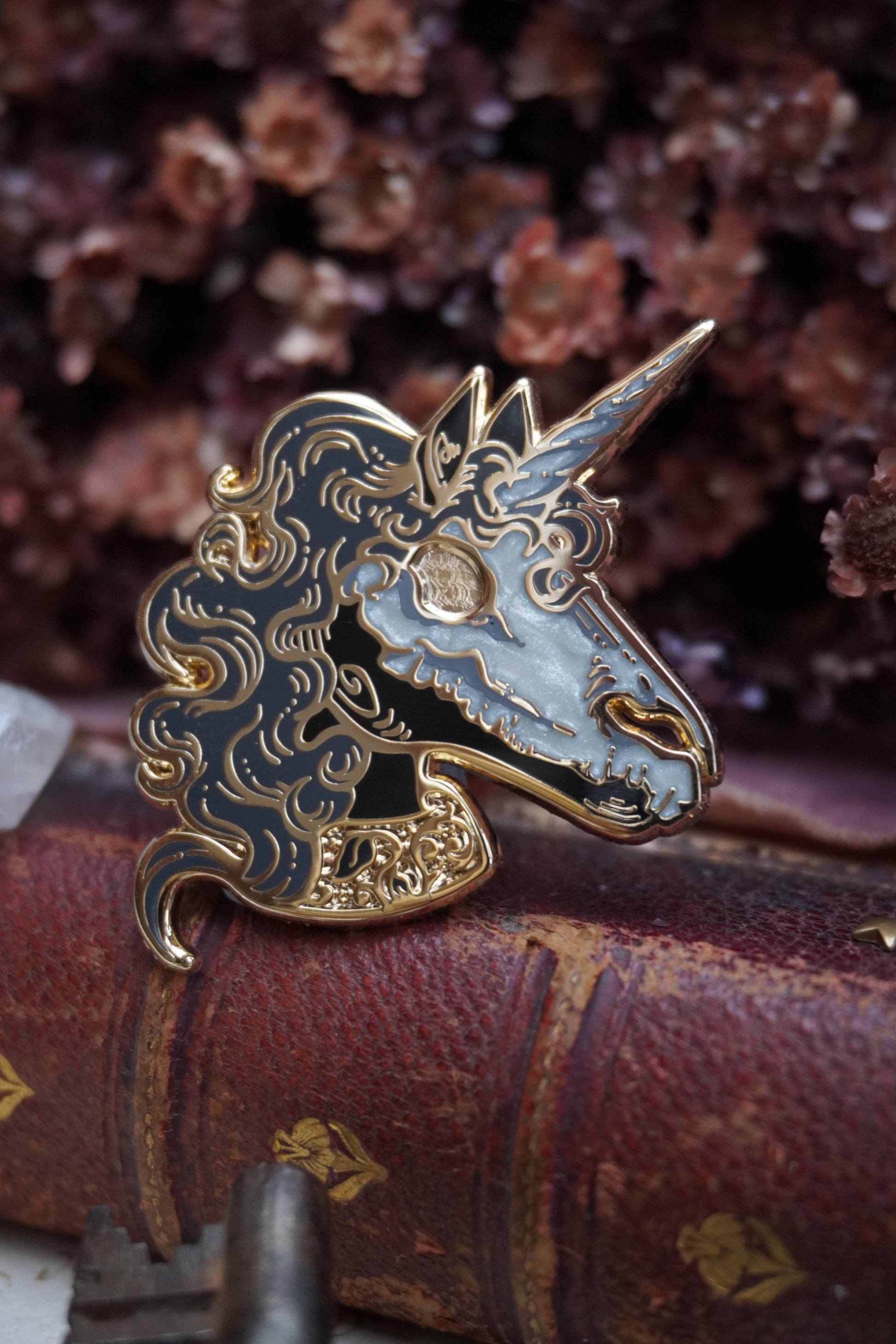 A close up of an enamel pin shaped like a unicorn head. The pin features a black face and neck with white pearl effect skull on top of the face, grey curly mane, lighter grey screen print details and gold plated metal outlines resting on vintage paper with dried dark red flowers in the background and blurred keys in front