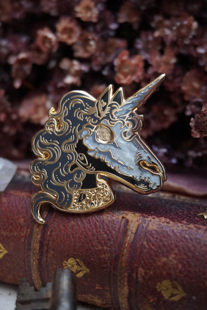 A close up of an enamel pin shaped like a unicorn head. The pin features a black face and neck with white pearl effect skull on top of the face, grey curly mane, lighter grey screen print details and gold plated metal outlines resting on vintage paper with dried dark red flowers in the background and blurred keys in front