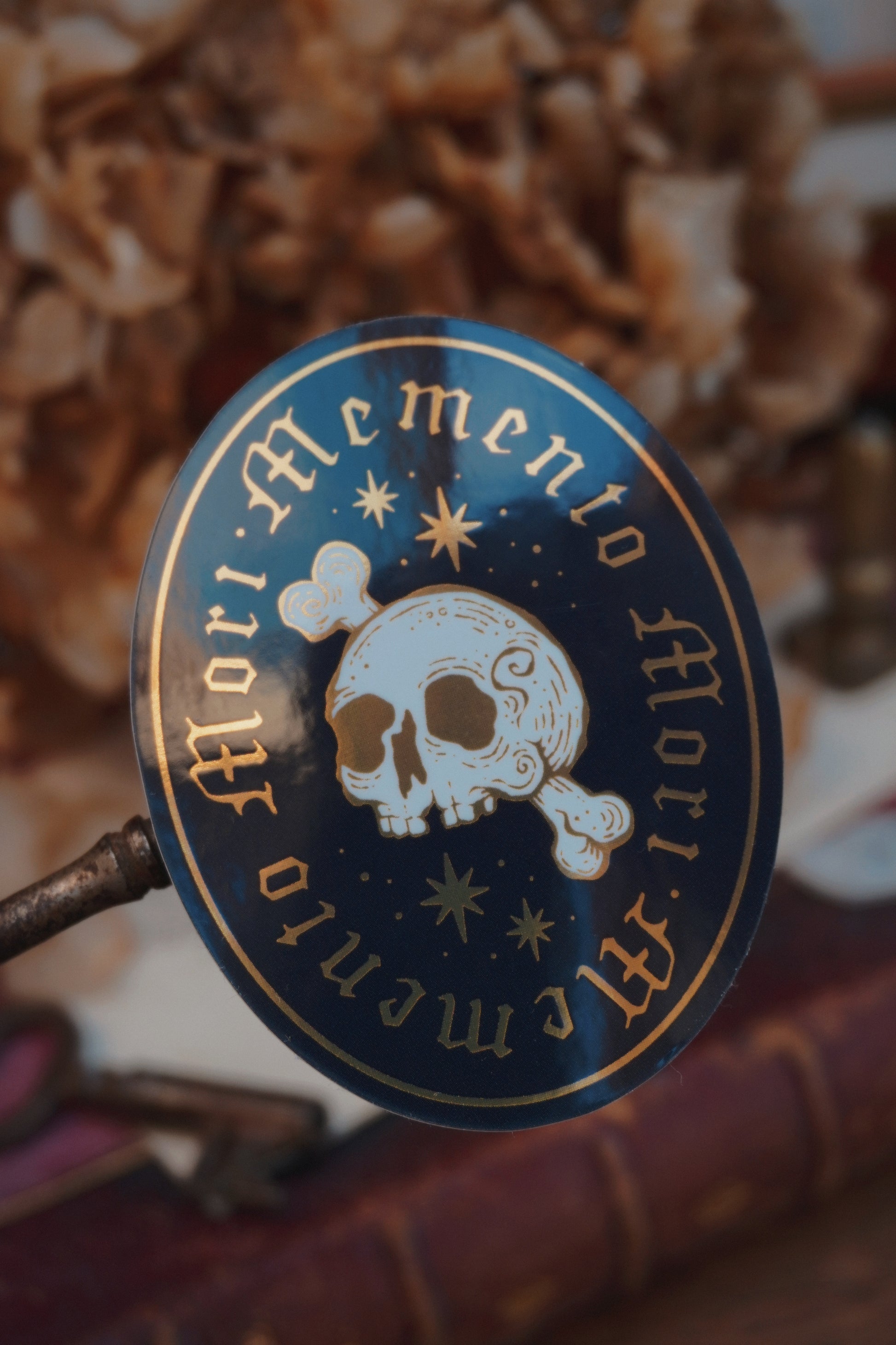 A close-up of an ovel sticker in white, black and gold, with gold lettering reading 'Memento Mori' and a white skull-and-crossbones motif, shown against a blurred background of dried flowers and books.