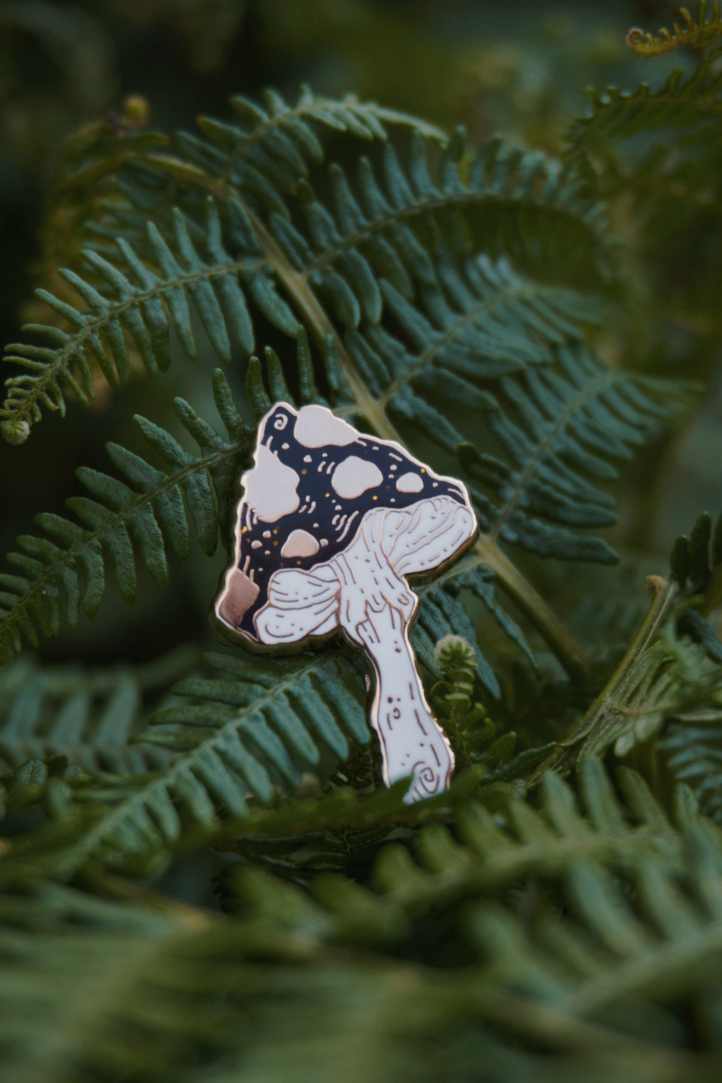 A mushroom shaped enamel pin with a black and gold spotted cap with gold glitter flecks and white stem, photographed resting on green fern leaves.