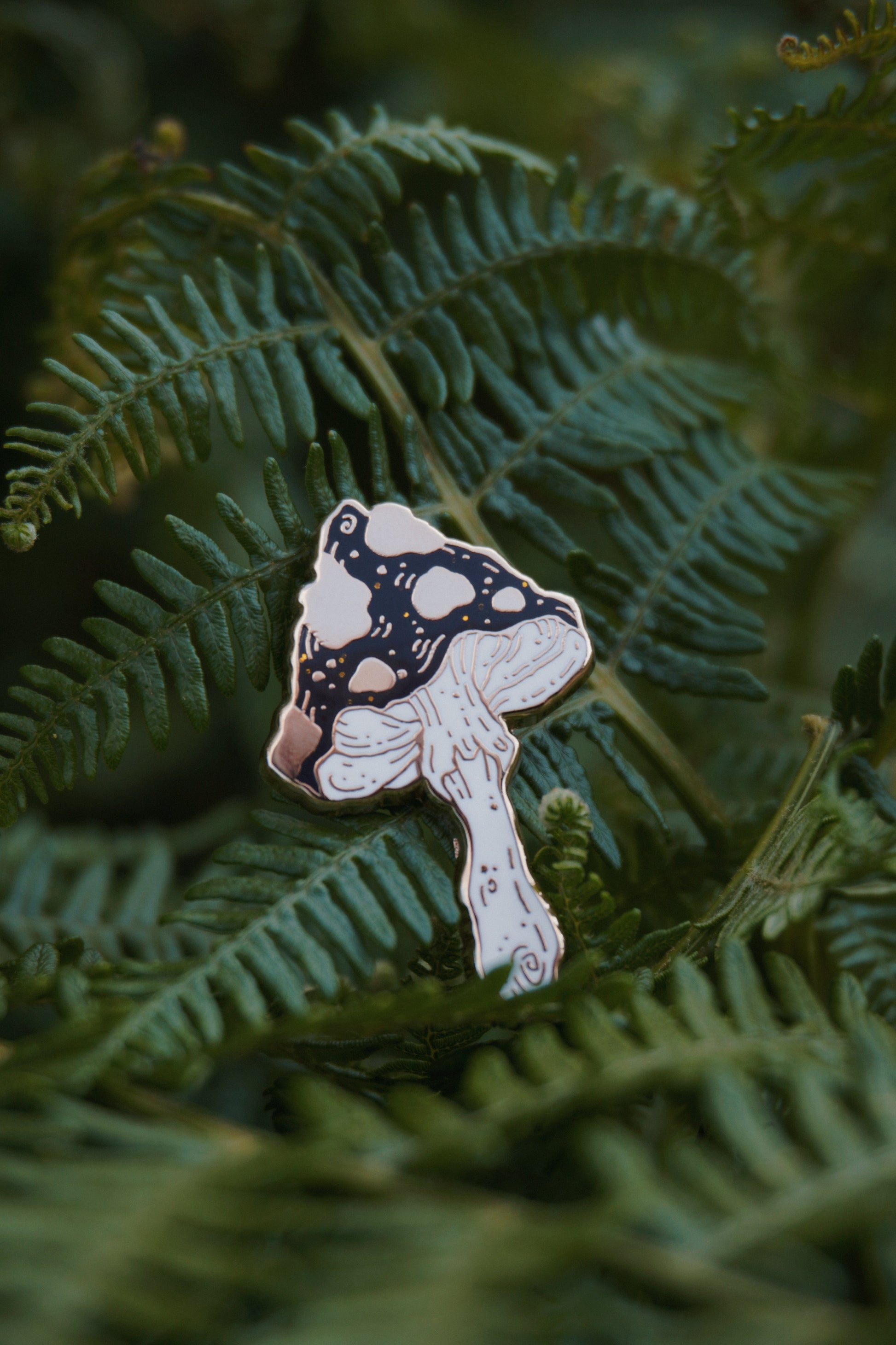 A mushroom shaped enamel pin with a black and gold spotted cap with gold glitter flecks and white stem, photographed resting on green fern leaves.