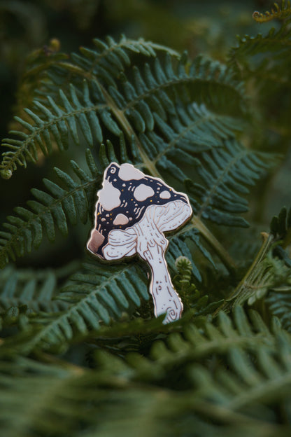 A mushroom shaped enamel pin with a black and gold spotted cap with gold glitter flecks and white stem, photographed resting on green fern leaves.
