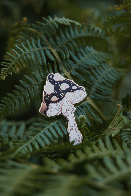 A mushroom shaped enamel pin with a black and gold spotted cap with gold glitter flecks and white stem, photographed resting on green fern leaves.
