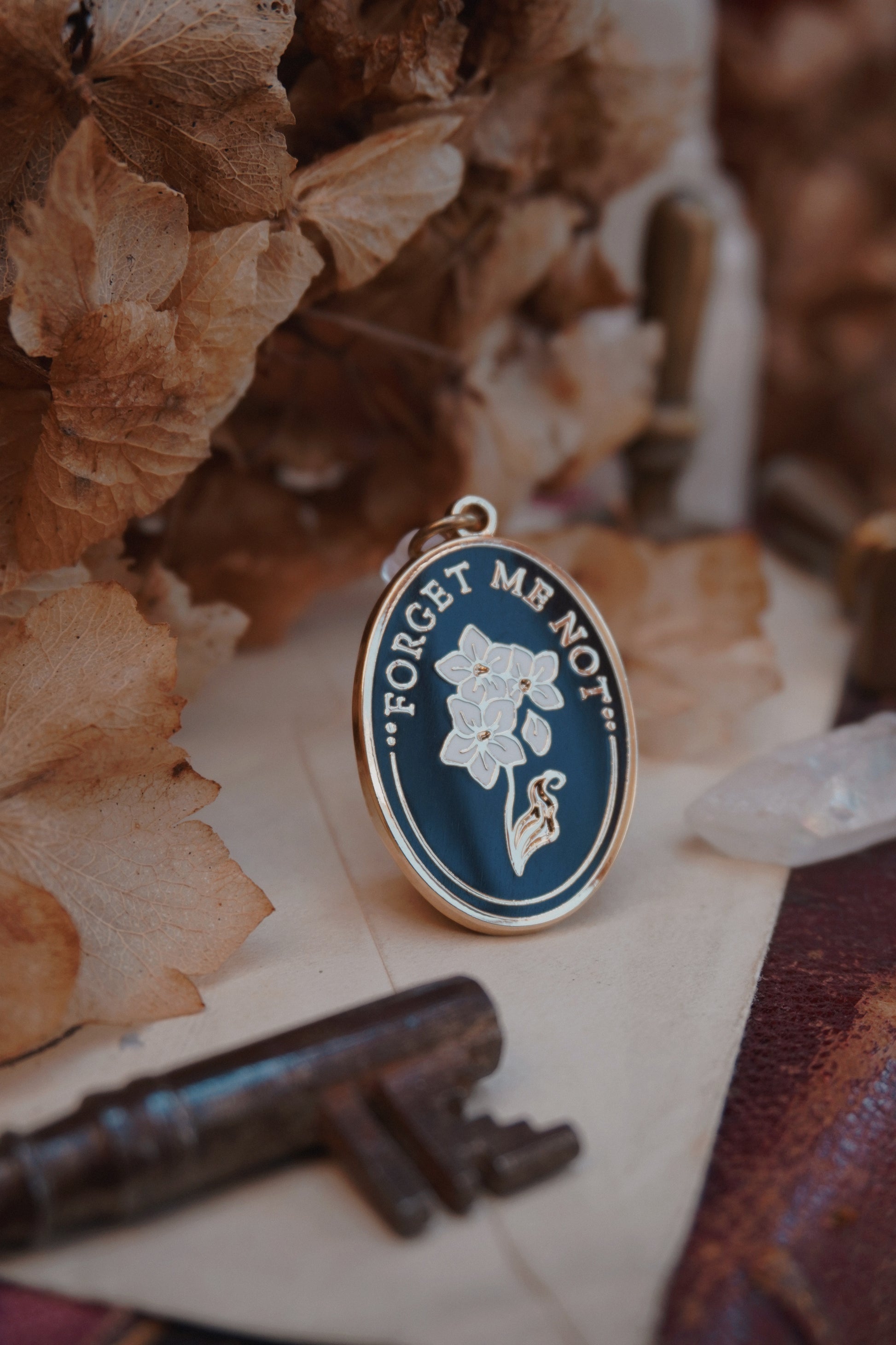 An oval enamel dust plug pendant black background, white floral design, and the text 'FORGET ME NOT' in gold, styled on vintage paper with dried flowers, a vintage key, and a crystal.