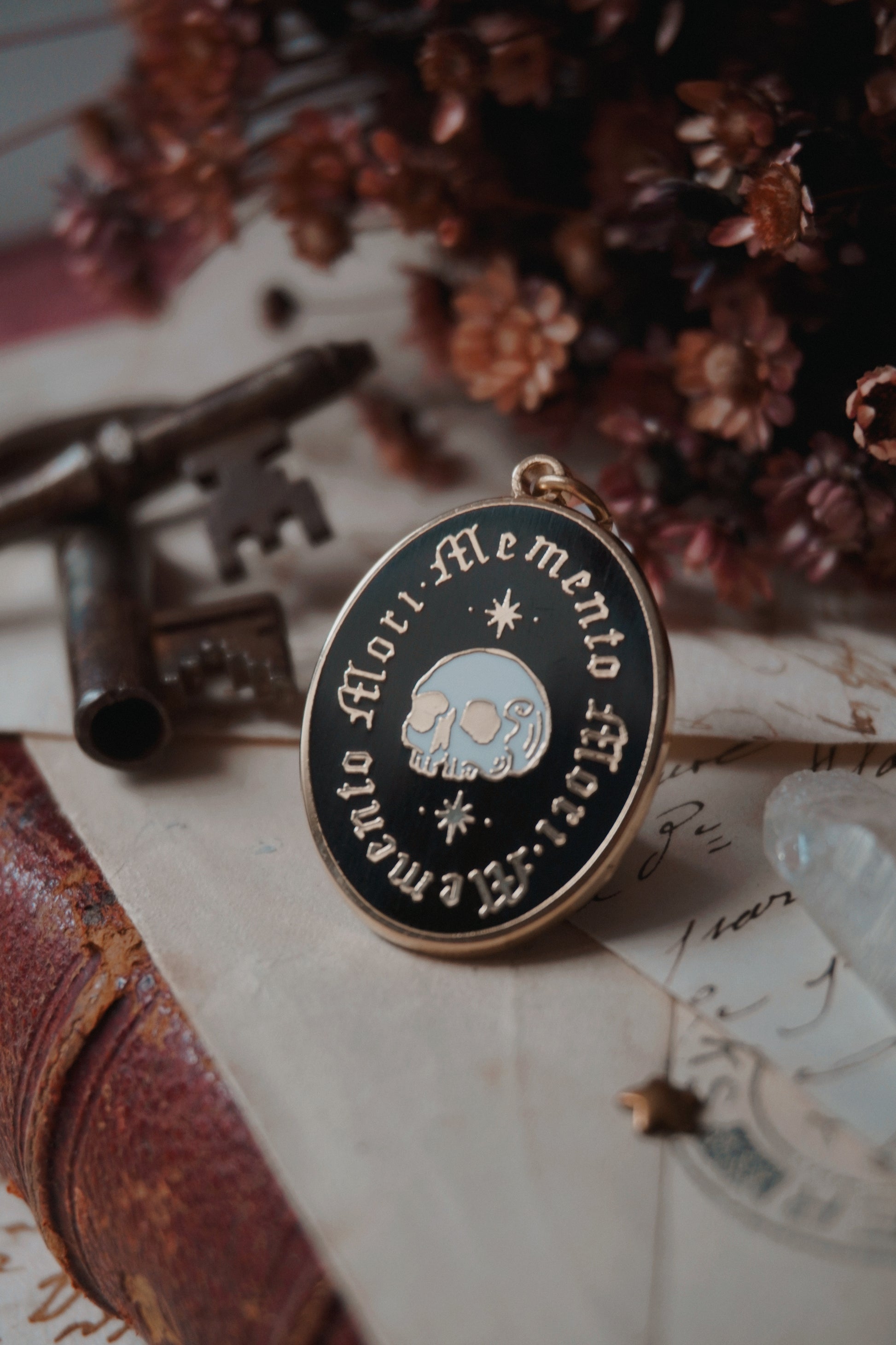 An oval enamel dust plug pendant black background, gold and white skull an stars, and the text 'Memento Mori' framed twice in gold, styled on vintage paper with dried flowers, a vintage key, and a crystal.