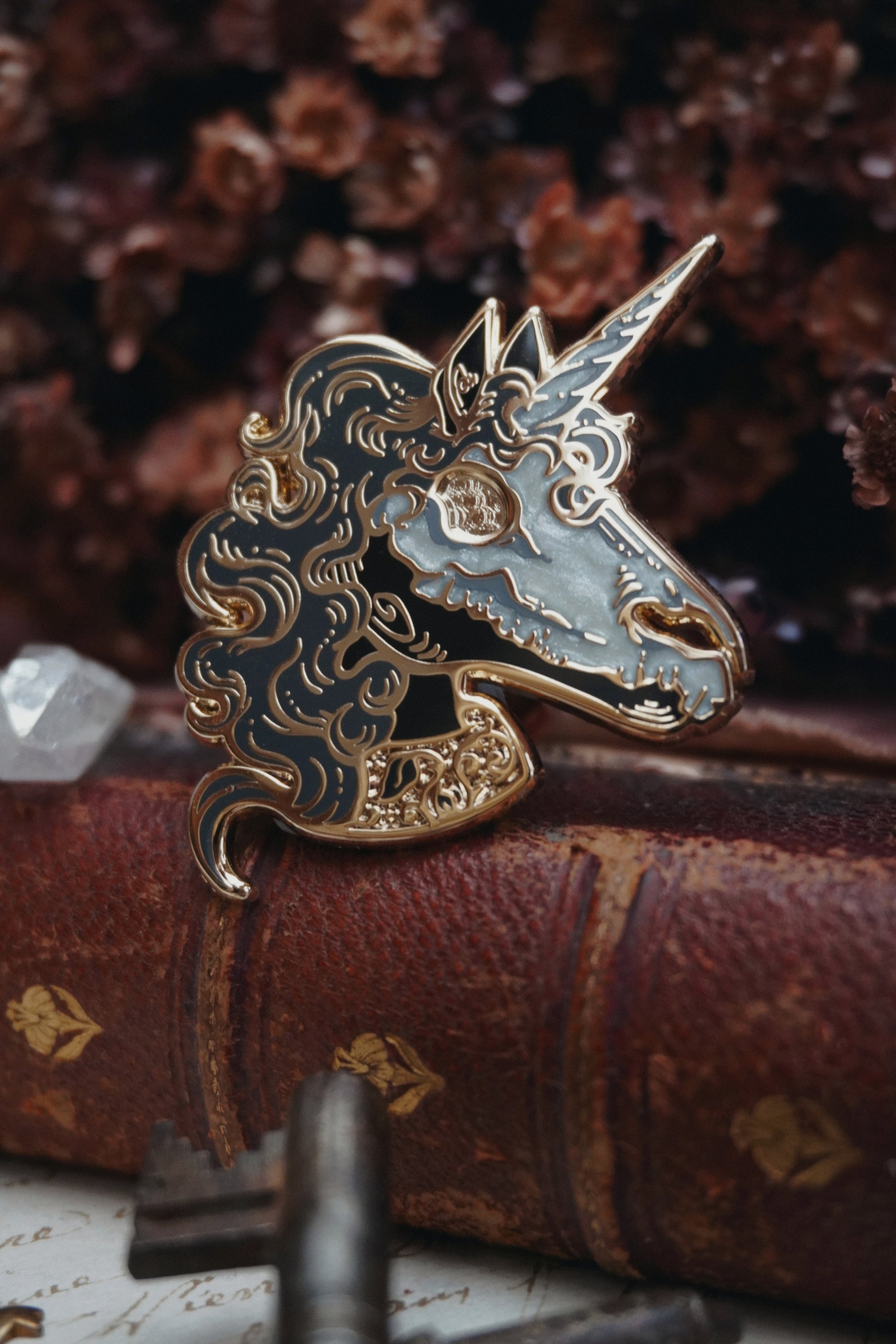 A close up of an enamel pin shaped like a unicorn head. The pin features a black face and neck with white pearl effect skull on top of the face, grey curly mane, lighter grey screen print details and gold plated metal outlines resting on vintage paper with dried dark red flowers in the background and blurred keys in front