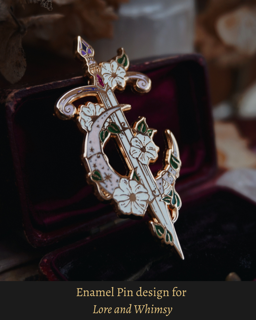 Ornate enamel pin displayed on a vintage dark red and velvet jewellery box. The pin is a gold metal outlined with a sword and moon/wrapping flowers design. The moon is pink pearl swirl with stars, green leaves have additional green screen print details with white glitter flowers and purple and pink details in the handle. 