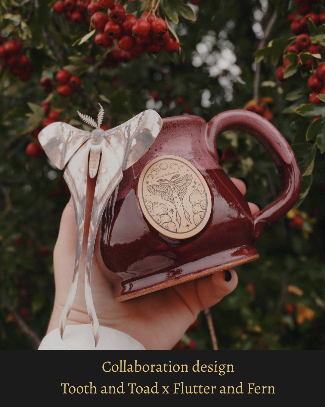 A red and pink stoneware mug with decorative medallion of a moth, clouds, moons and stars, held in a hand, surrounded by red berries and green leaves. Contains text that states it is a: 'Collaboration design Tooth and Toad x Flutter and Fern'.