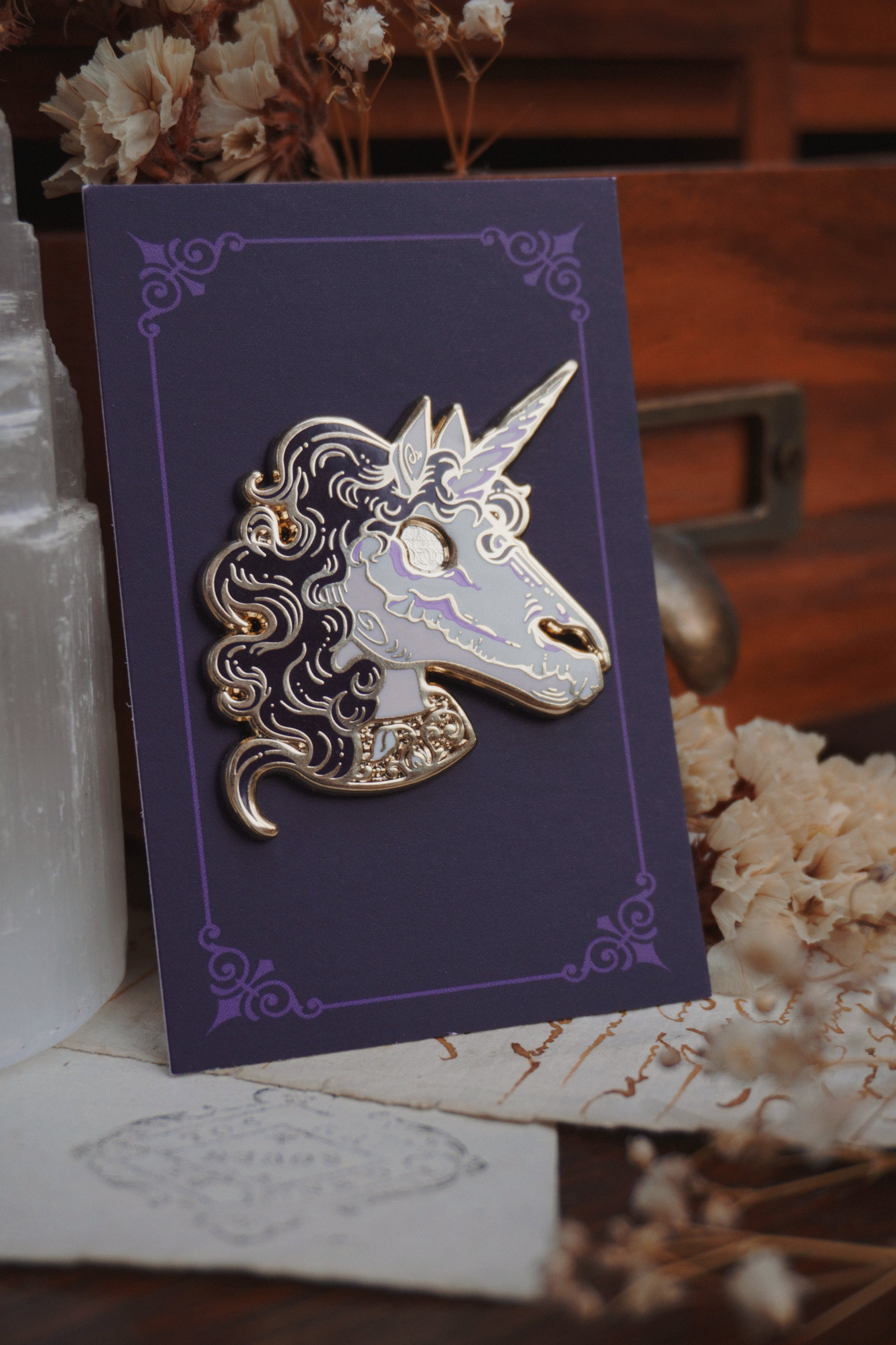 An undead unicorn enamel pin on a purple backing card with a decorative border