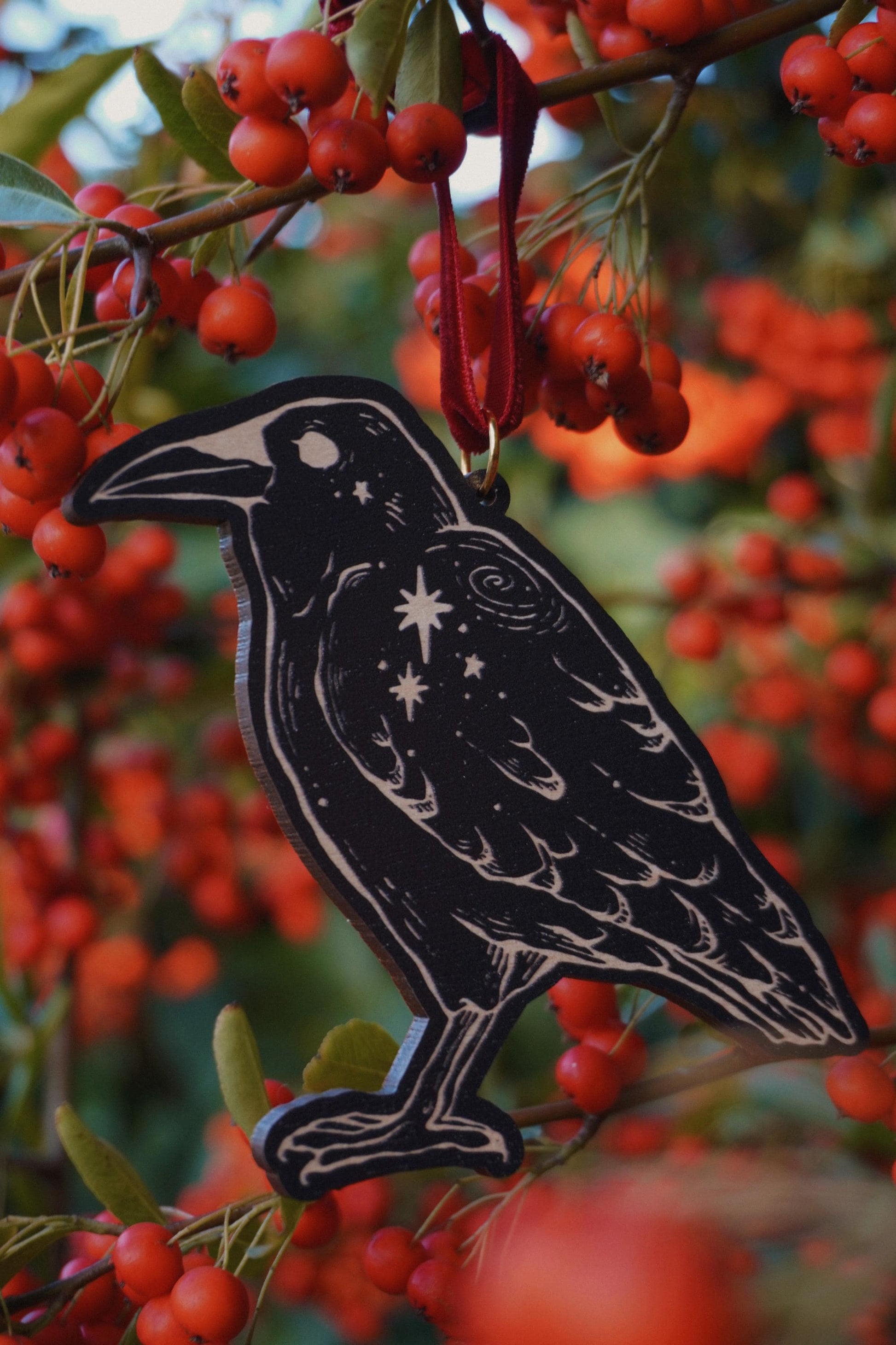 Wooden hanging ornament in the shape of a crow with a crow and stars design, resting on a red berry bush